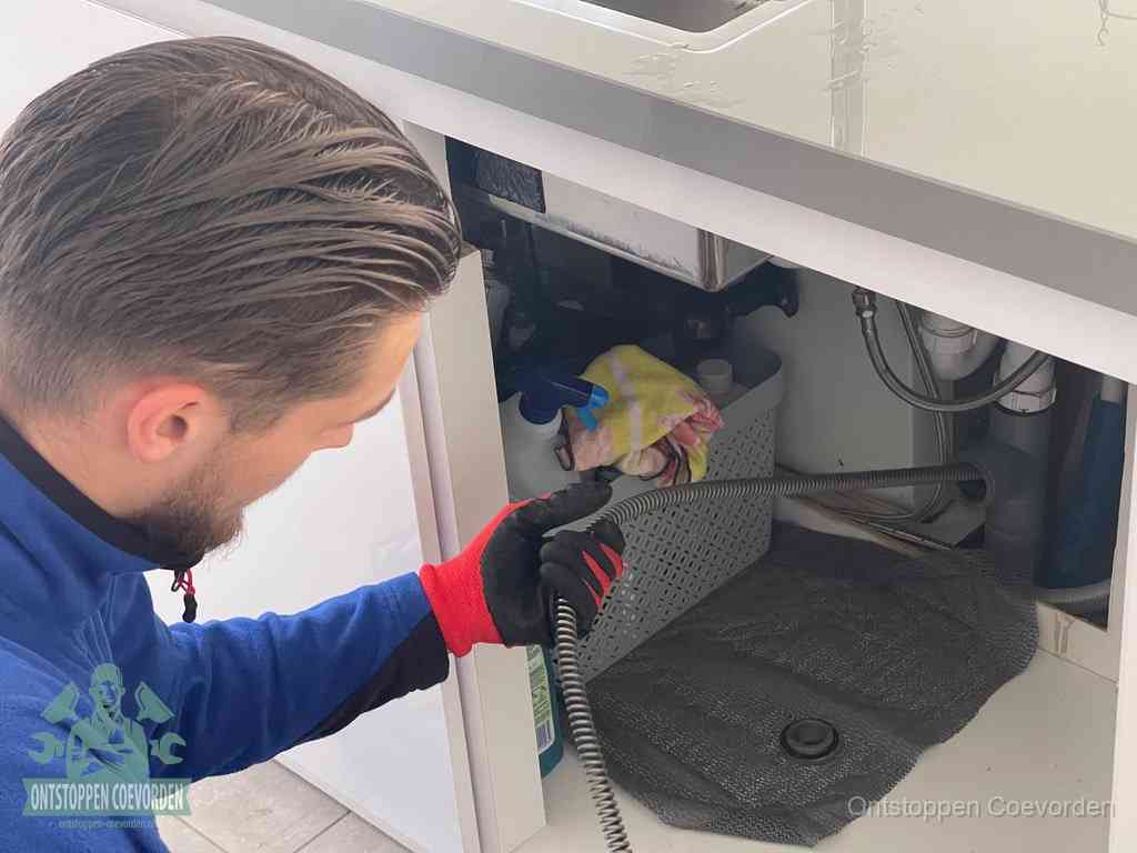 Een monteur voert een flexibele ontstoppingsveer in onder een keukenkastje om een verstopping voorbij de sifon te bereiken in Coevorden