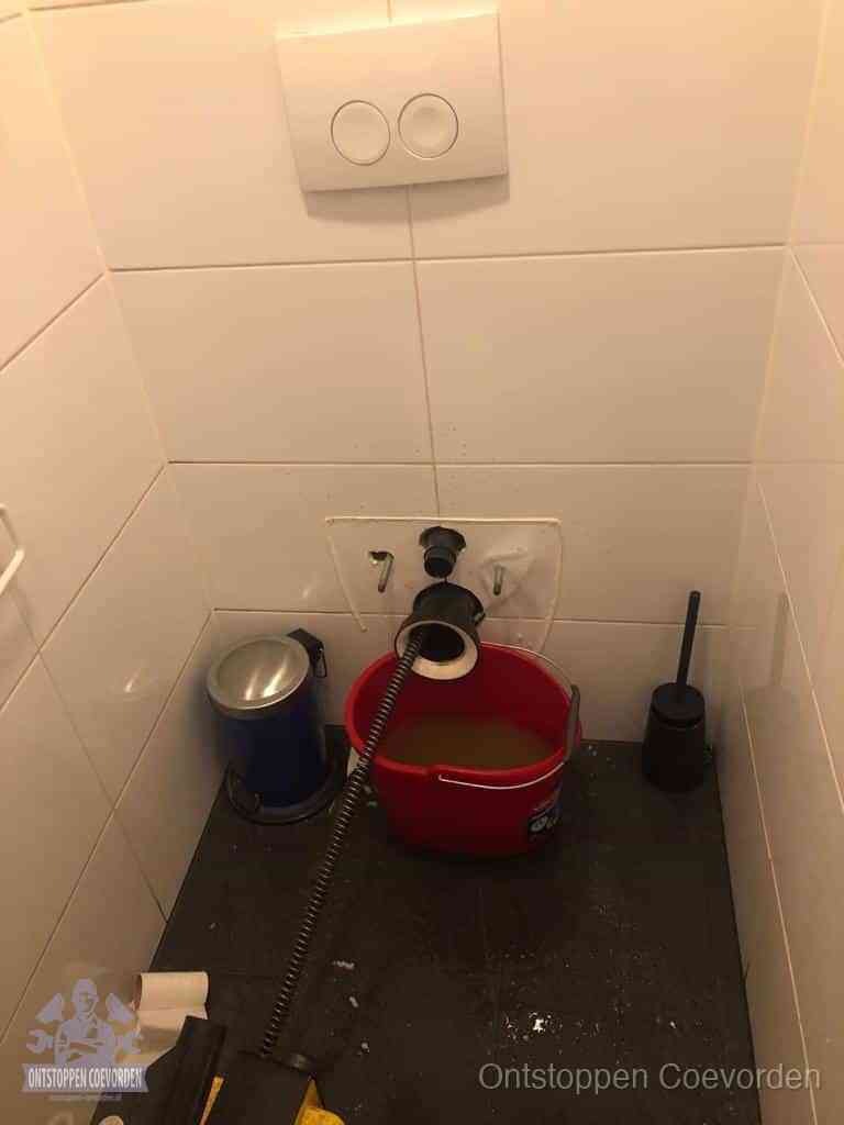 Een ontstopte wand-wc met verwijderde toiletpot waar een ontstoppingsveer in de wanduitgang verdwijnt, het achterhalen waar de verstopping zit in Coevorden