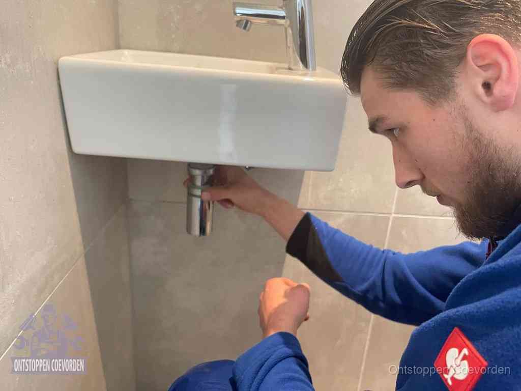 Een monteur draait met de hand de zwanenhalssifon onder een fonteinwasbak in de toiletruimte los om de bron van rioollucht in Coevorden te vinden