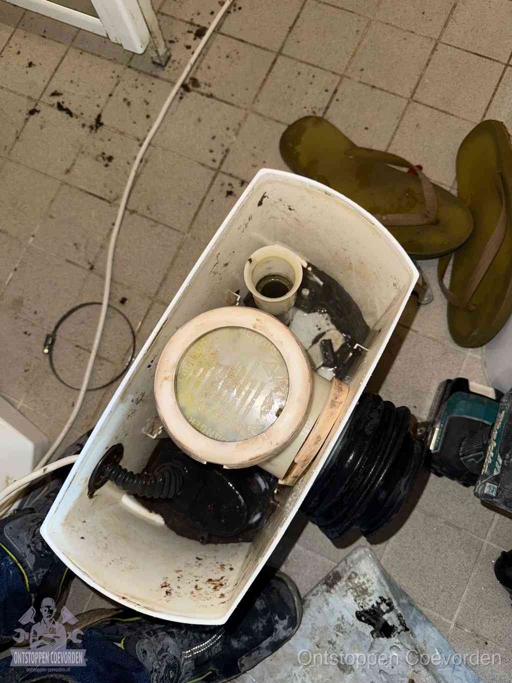 Een gedemonteerde toiletpot op de vloer met ontstoppingsveer en gereedschap ernaast, het verwijderen van een vastzittend voorwerp uit de wc in Coevorden